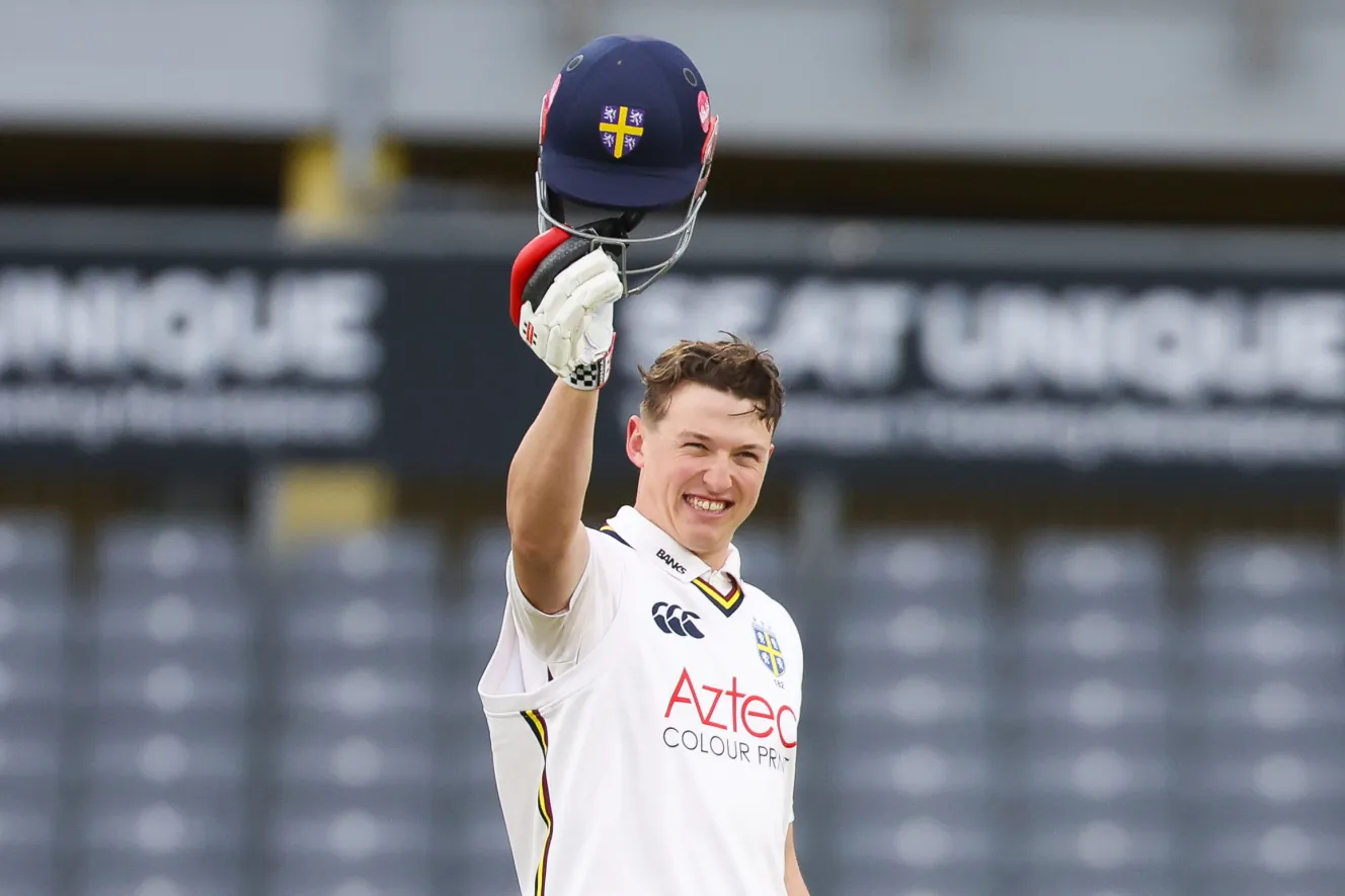Ben McKinney of Durham celebrates his double hundred in a white cricket uniform, holding his dark blue helmet up.