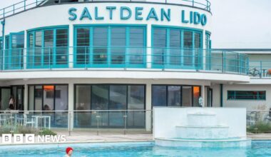 A white and turquoise Art Deco lido, with Saltdean Lido written in lettering across its frontage. A woman in a red bathing cap swims in the pool in front of it.