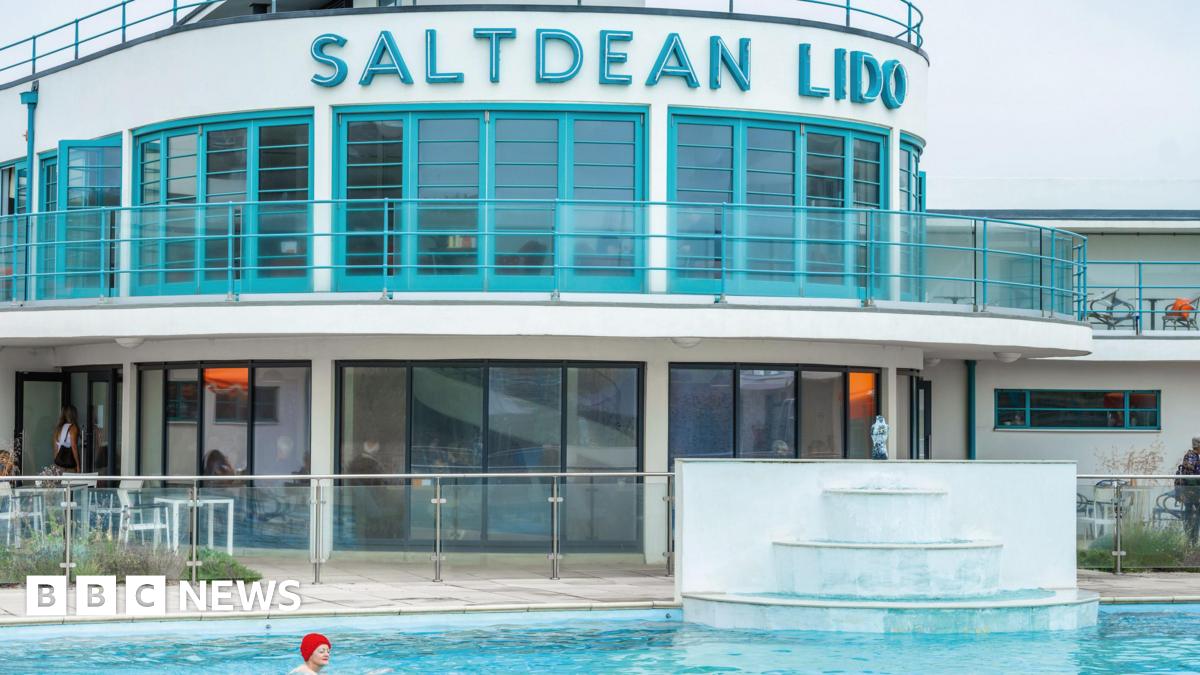 A white and turquoise Art Deco lido, with Saltdean Lido written in lettering across its frontage. A woman in a red bathing cap swims in the pool in front of it.