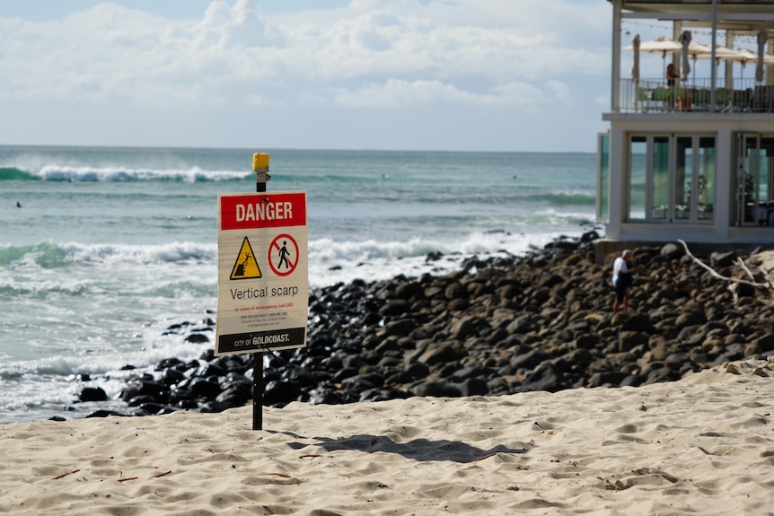 A warning sign is visible on a sandy beach with rocks in the shallows in the background.