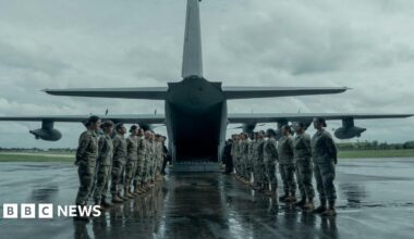 Military personnel form two lines, facing each other, to the rear of a military aircraft which has its ramp down at the bottom. It is a grey and wet day. The servicemen and women have their hands placed behind their backs.