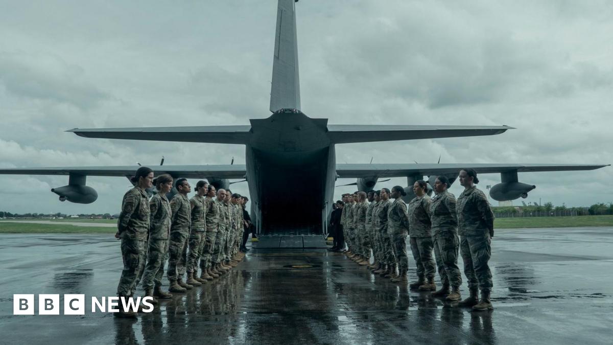 Military personnel form two lines, facing each other, to the rear of a military aircraft which has its ramp down at the bottom. It is a grey and wet day. The servicemen and women have their hands placed behind their backs.