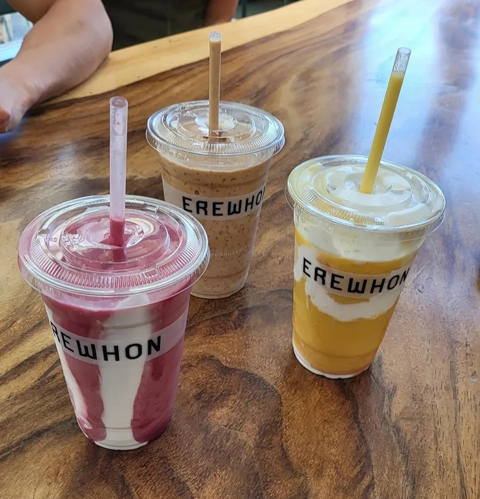 Three smoothies on a wooden table, each in a cup labeled "Erewhon." They are strawberry, mocha, and mango flavors from left to right