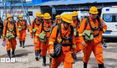 Rescuers in orange safety suits and helmets walking near industrial buildings