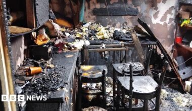 A room is charred and blackened by fire. Cupboard doors are black from fire damage. Chairs are broken and charred. Insulation is lying on the counter tops. Paint has been stripped from the walls.