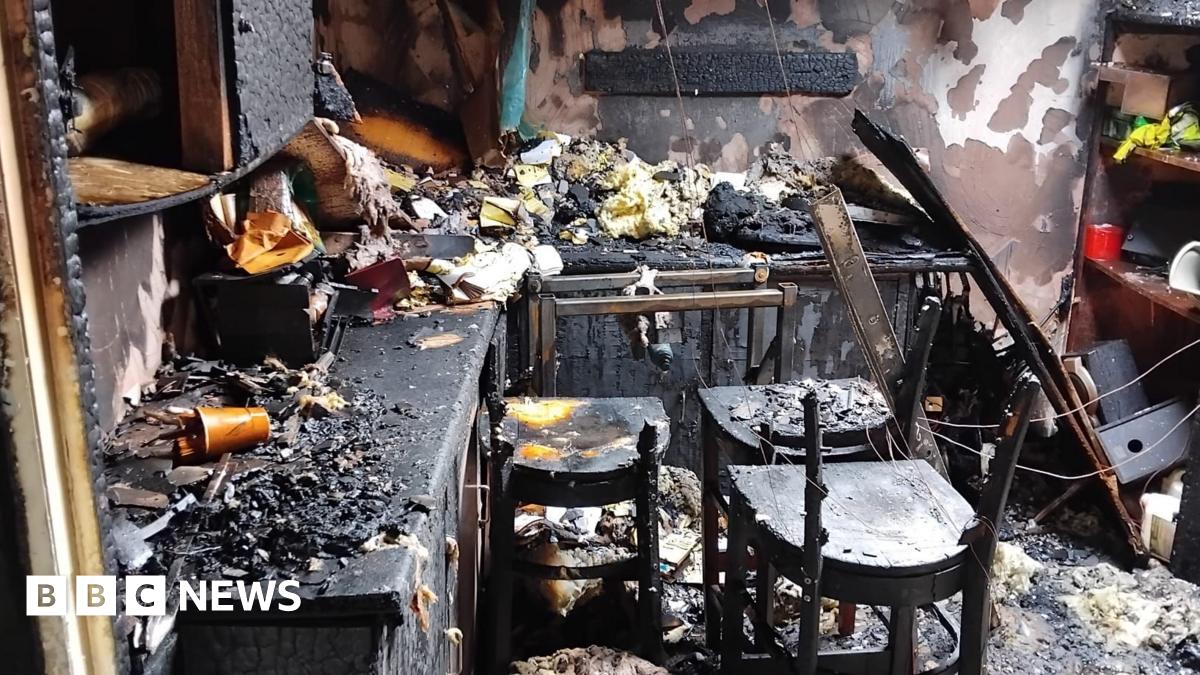 A room is charred and blackened by fire. Cupboard doors are black from fire damage. Chairs are broken and charred. Insulation is lying on the counter tops. Paint has been stripped from the walls.