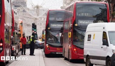 A busy central London street with multiple red double-decker buses stuck in slow-moving traffic. A white van and several cars are also queued, while two men stand on the pavement beside a bus. Christmas lights hang above the road, and buildings line the background.