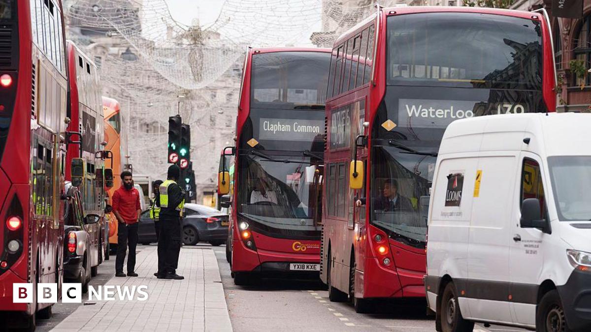 A busy central London street with multiple red double-decker buses stuck in slow-moving traffic. A white van and several cars are also queued, while two men stand on the pavement beside a bus. Christmas lights hang above the road, and buildings line the background.