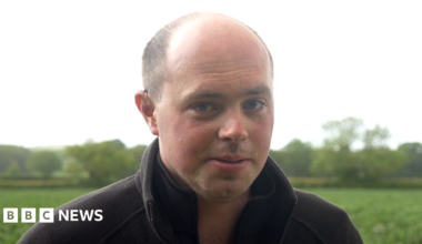 Matthew Register has a shaved head and is wearing a black jumper in front of a green field.