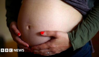 A close up of a woman cradling her pregnant belly. She is wearing red nail varnish and a silver ring.