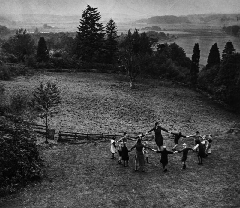A group of children and an adult stand in a circle holding hands on a grassy field surrounded by trees, with a scenic landscape and hills visible in the background.