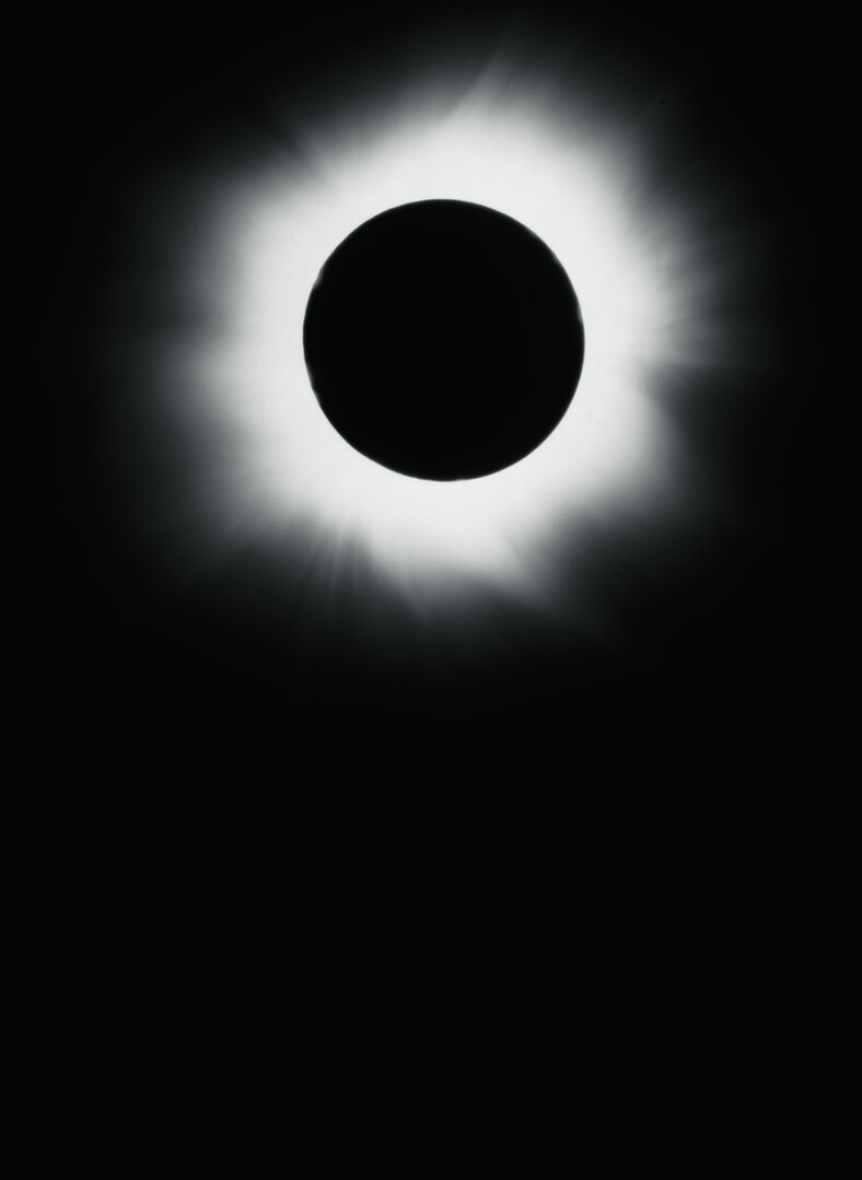 A stark, vertical black and white photograph of a total solar eclipse, showing a perfectly dark lunar disk centered within a glowing, radiant white solar corona against an absolute black sky.