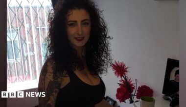 A woman in her 40s, with curly blackhair and wearing a black vest top, smiles at the camera by an upstairs window. She has a sleeve of black and white tattoos on her right arm including an image of a lighthouse and a woman.