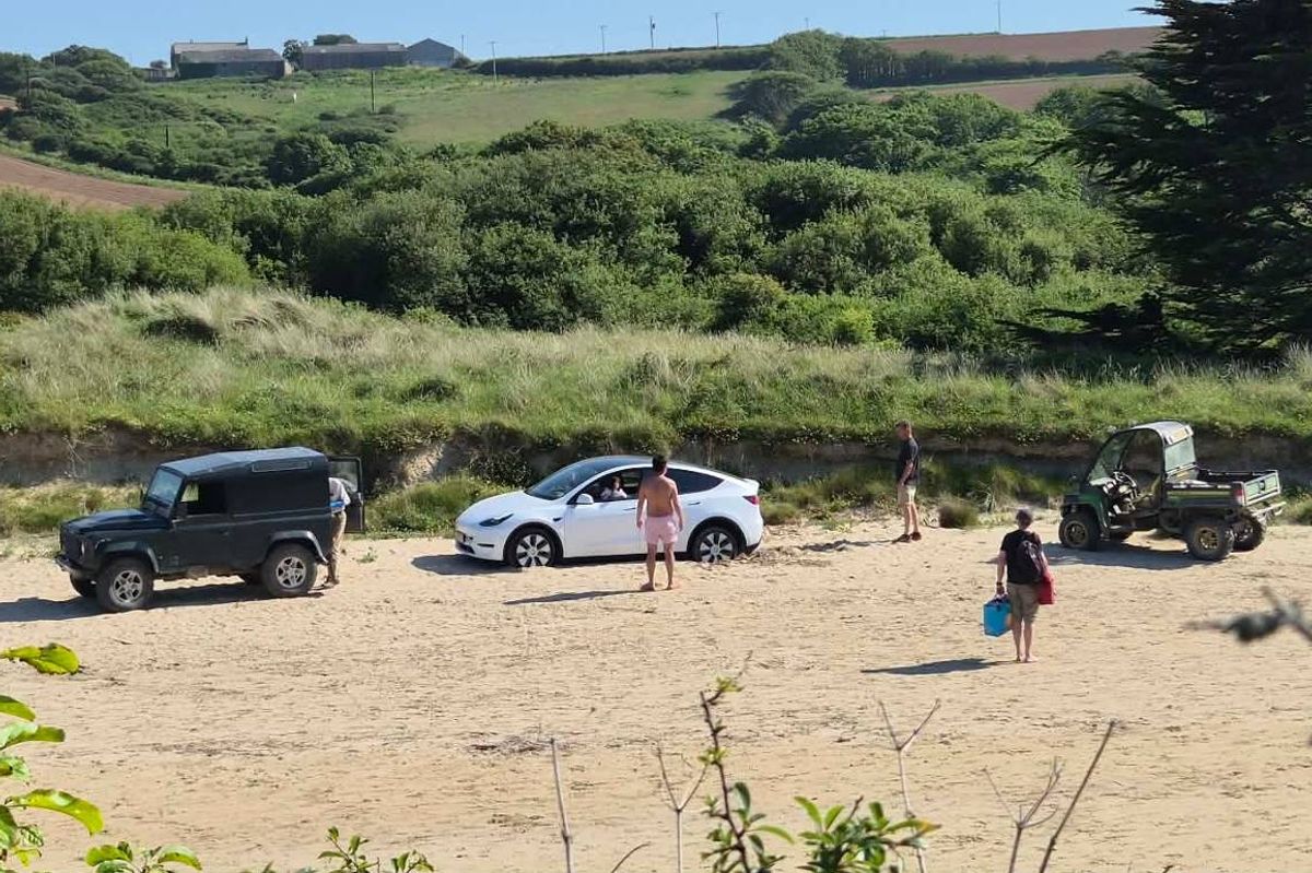 An outdoor scene depicts several vehicles parked in a sandy area. A small group of individuals can be seen walking around, some of whom are carrying bags. The surrounding landscape is characterized by lush greenery and bushes, with a clear sky overhead.