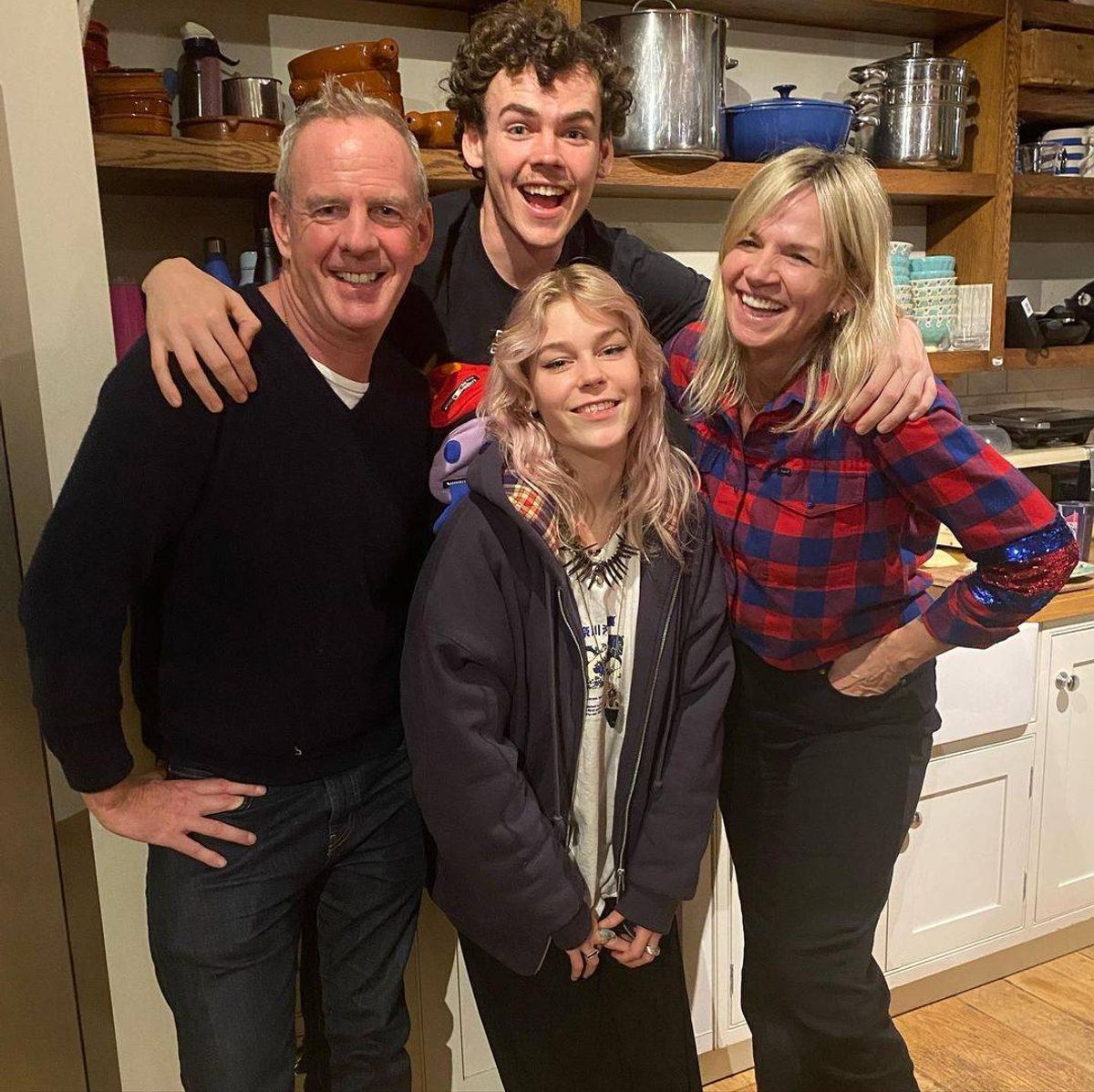 A family of four is gathered together in a kitchen, with two women standing on either side of two men. The two men have their arms around the shoulders of the women, with one man’s arm around the woman on his right, and the other man’s arm around the woman on his left. The family is smiling and posing for a photograph, with shelves in the background.