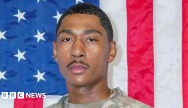 close-up of a soldier's face, looking serious and wearing a camo uniform, with an American flag int he background