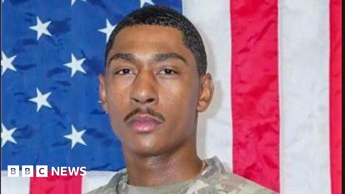 close-up of a soldier's face, looking serious and wearing a camo uniform, with an American flag int he background