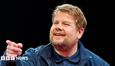 James Corden has sandy blonde hair and a beard and moustache. He is wearing a blue jacket, blue polo shirt and is pointing off to the side of the camera.