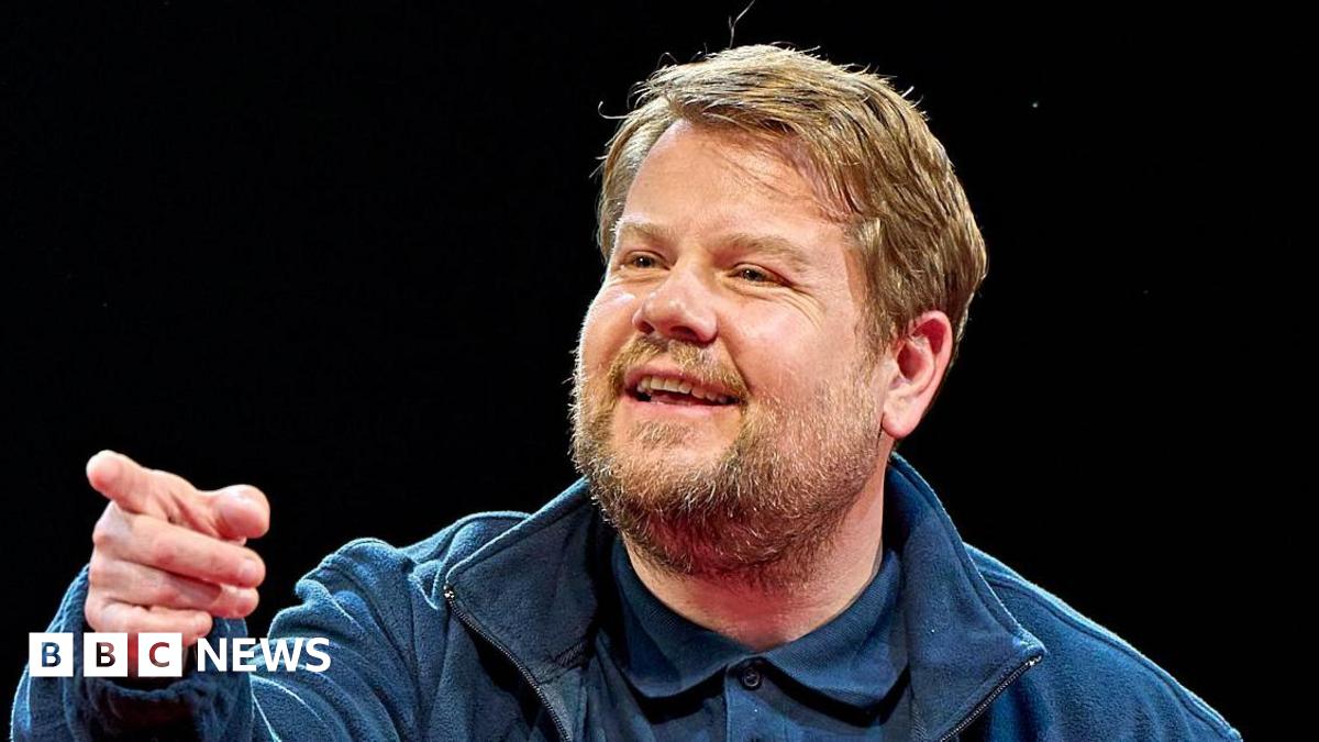 James Corden has sandy blonde hair and a beard and moustache. He is wearing a blue jacket, blue polo shirt and is pointing off to the side of the camera.