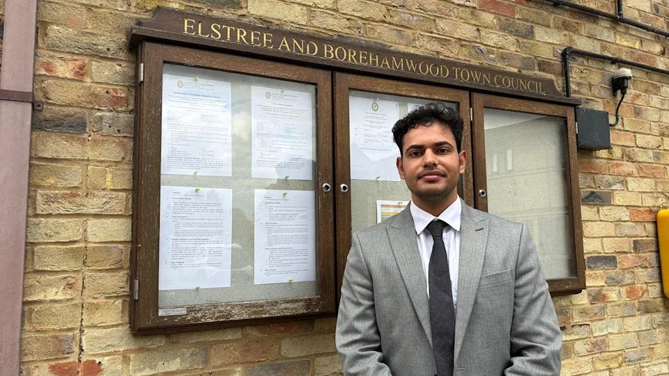 Tushar Kumar in a grey suit stands beside a noticeboard outside Elstree and Borehamwood Town Council, set against a light brick wall. The board is filled with official printed documents pinned neatly in rows. The formal setting and smart attire give the image a composed, civic feel.