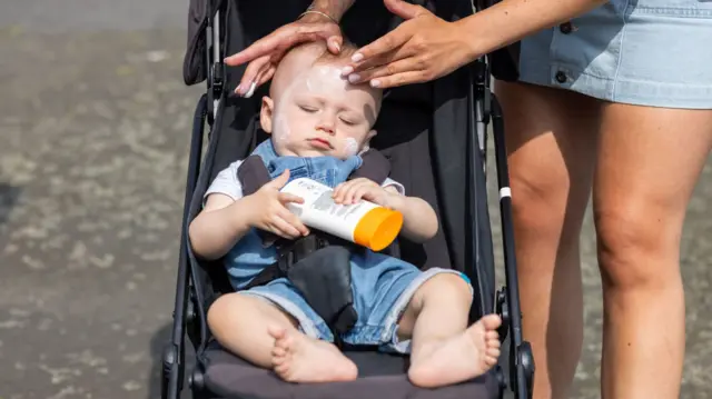 A baby lounges in a stroller, holding a sunscreen bottle while a caregiver gently applies lotion to his face