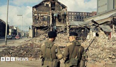 Two soldiers in green uniforms with their backs to camera, a bomb building is visible in front of them with debris all across the road