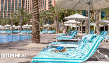 Blue-striped sun loungers covered in towels are lined up alongside a turquoise hotel swimming pool.