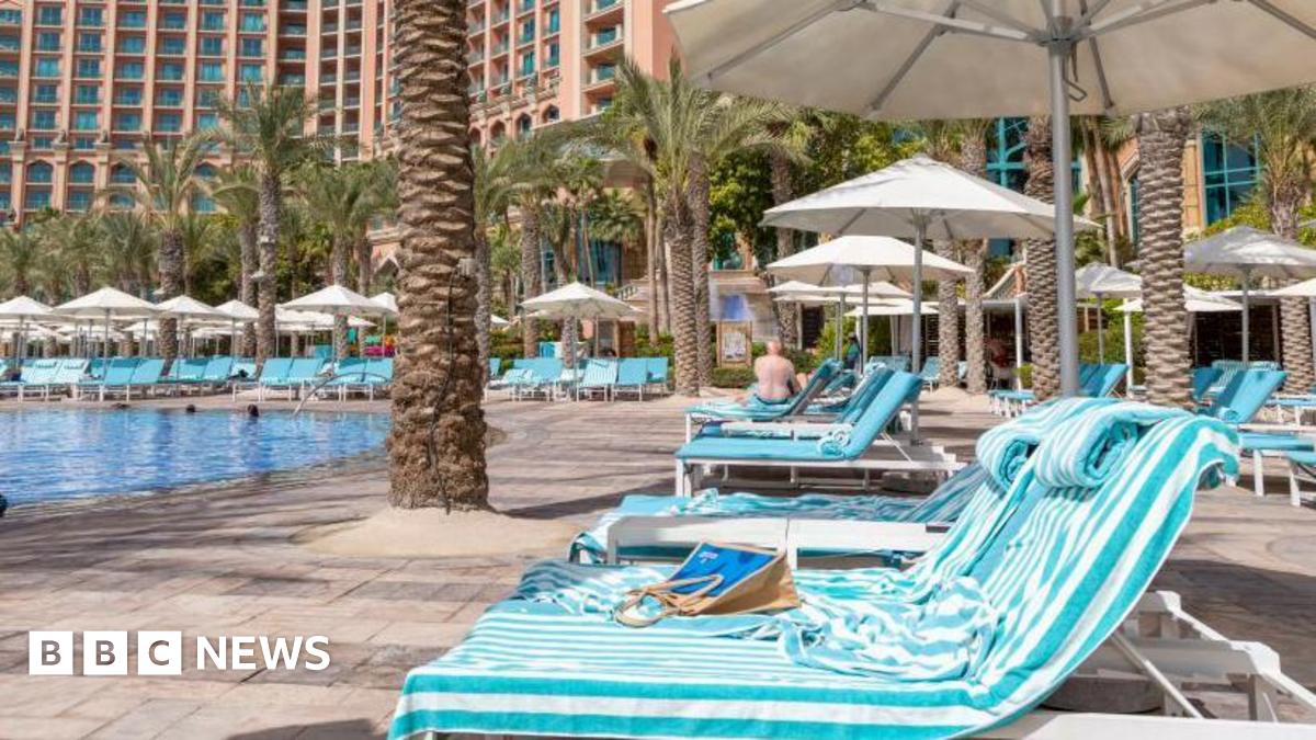 Blue-striped sun loungers covered in towels are lined up alongside a turquoise hotel swimming pool.