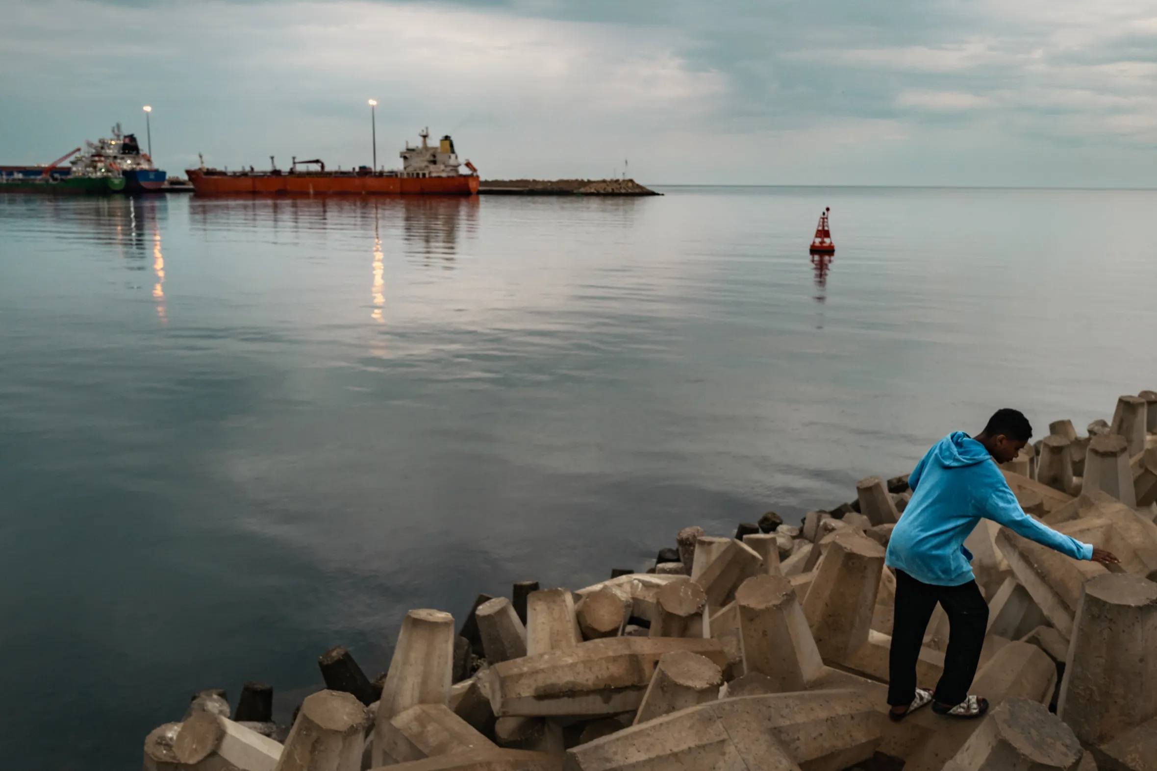 A bulk carrier anchored at Sultan Qaboos Port with a young man walking along concrete barriers in the foreground.