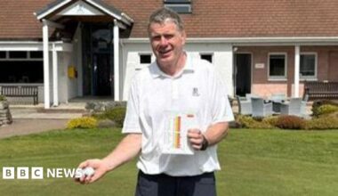 Douglas, stood on the grass outside the entrance to the Boat of Garten Golf Club. He is a man of average build with grey hair and is smiling. He's holding up two yellow flags and smiling.