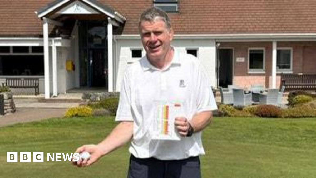 Douglas, stood on the grass outside the entrance to the Boat of Garten Golf Club. He is a man of average build with grey hair and is smiling. He's holding up two yellow flags and smiling.