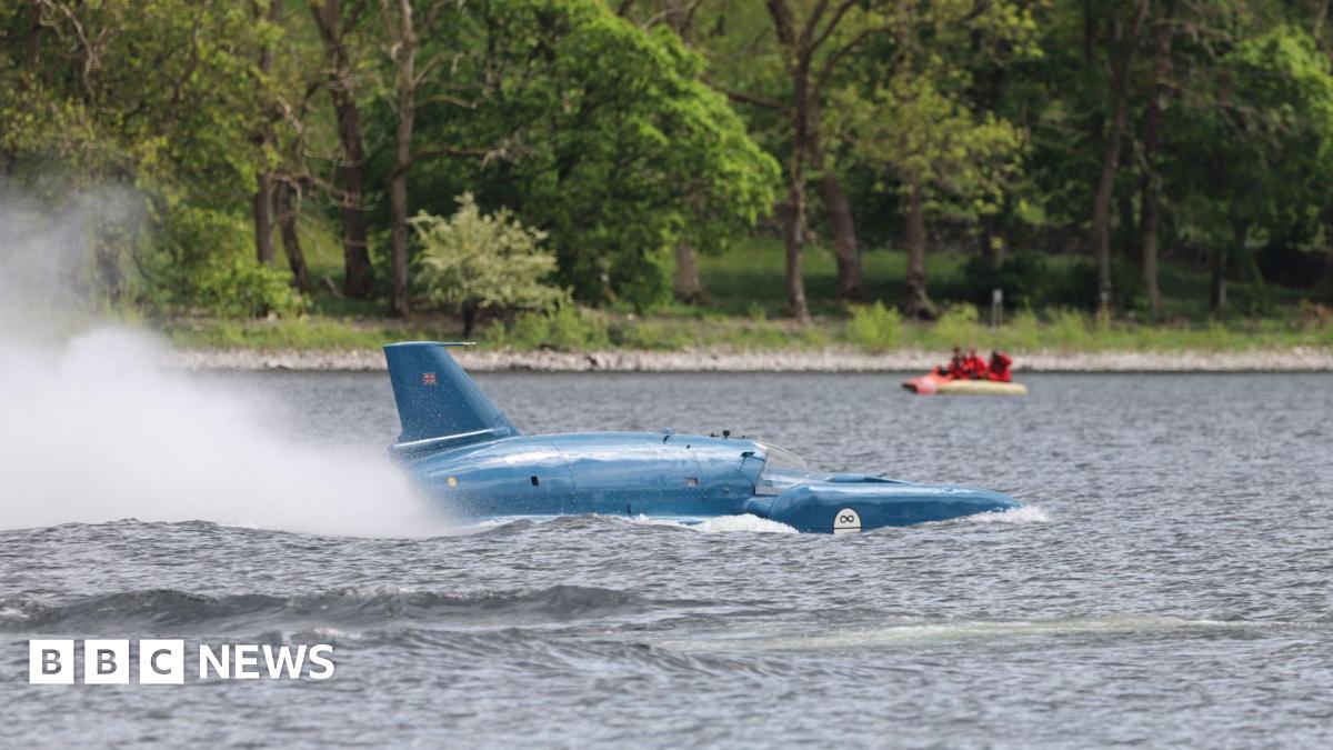A plume of spray rises from the back of Bluebird as it travels across Coniston Water. The machine looks like a cross between a boat and a plane. Painted blue, it has a tail fin. Trees can be seen on the shoreline.