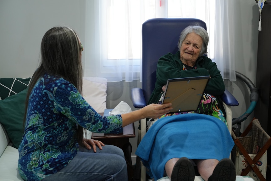 An elderly woman looks at a photo of herself as a younger person, while speaking to her daughter.