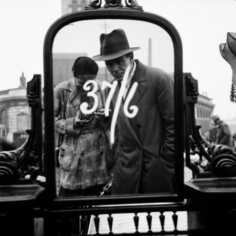 A black-and-white photo shows two people in coats and hats reflected in an ornate mirror with “37 1/6” written on the glass. The scene appears to be outdoors at a market with buildings in the background.