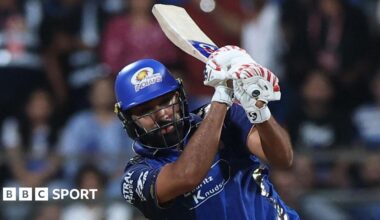 Rohit Sharma, wearing a blue shirt, blue helmet, and white batting gloves with red markings, holds his bat over his left shoulder after playing a shot