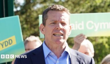 The image shows Rhun ap Iorwerth standing in front of people holding Plaid Cymru placards. He is wearing a navy suit and is mid-speech.
