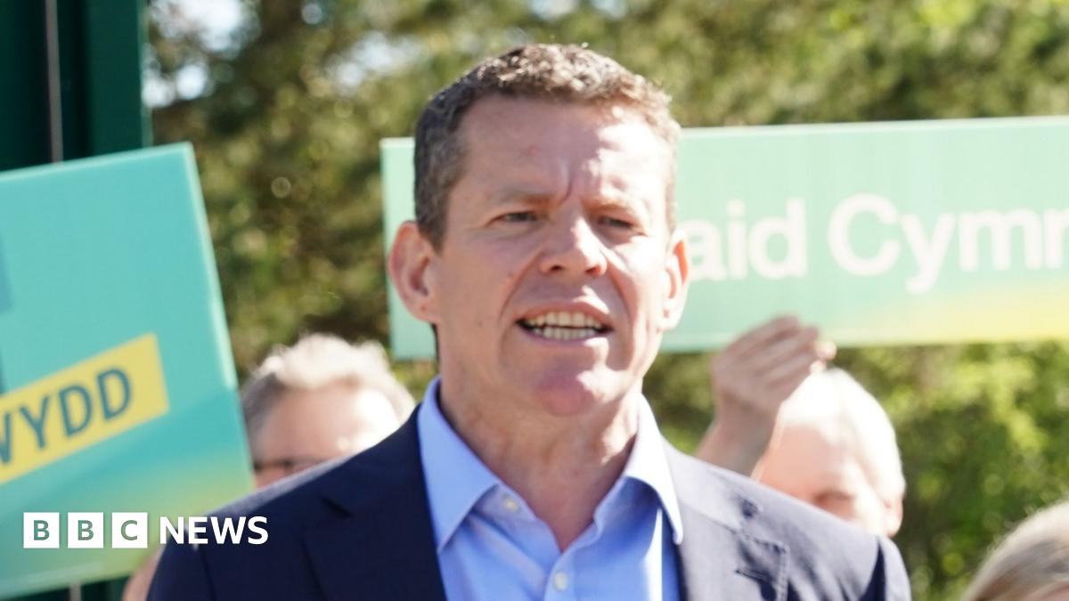 The image shows Rhun ap Iorwerth standing in front of people holding Plaid Cymru placards. He is wearing a navy suit and is mid-speech.