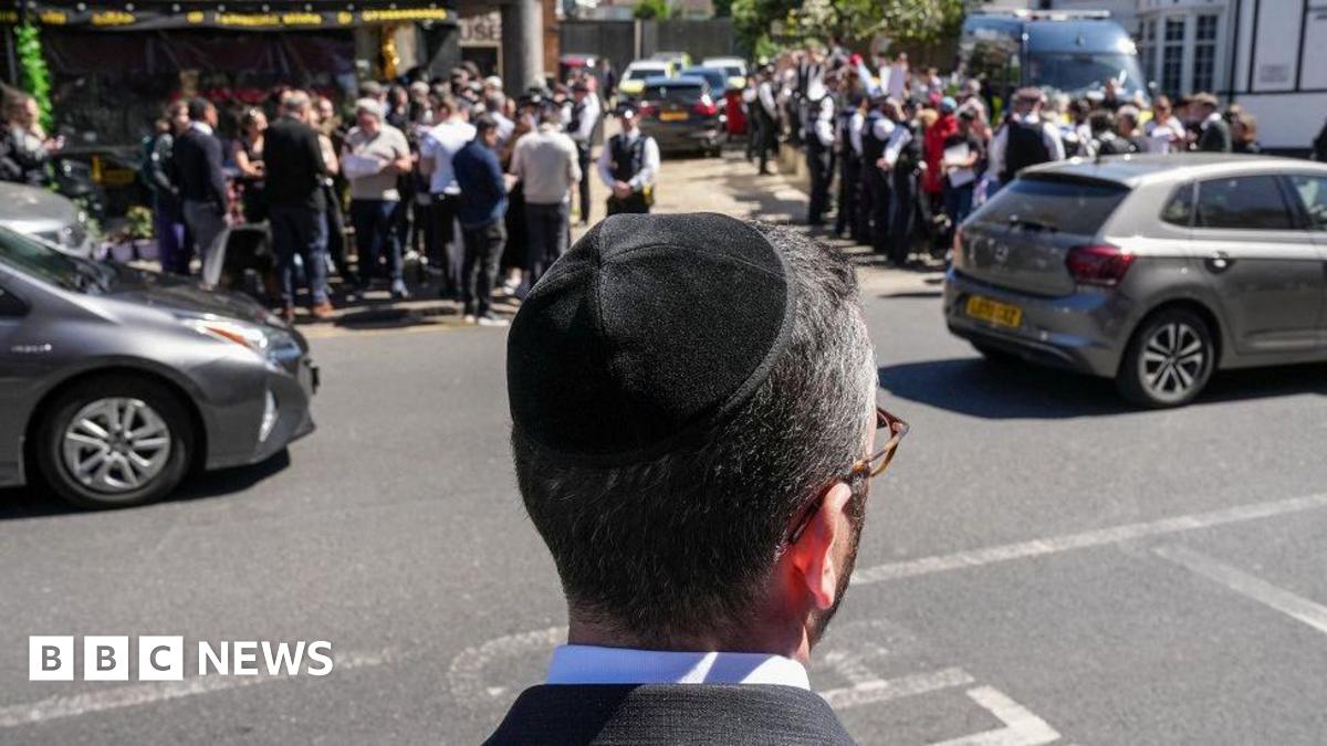 A Jewish man wearing a kippah is seen from behind as he stands looking out at a road as two cars pass in Golders Green. On the opposite pavement crowds are gathered with police officers nearby.