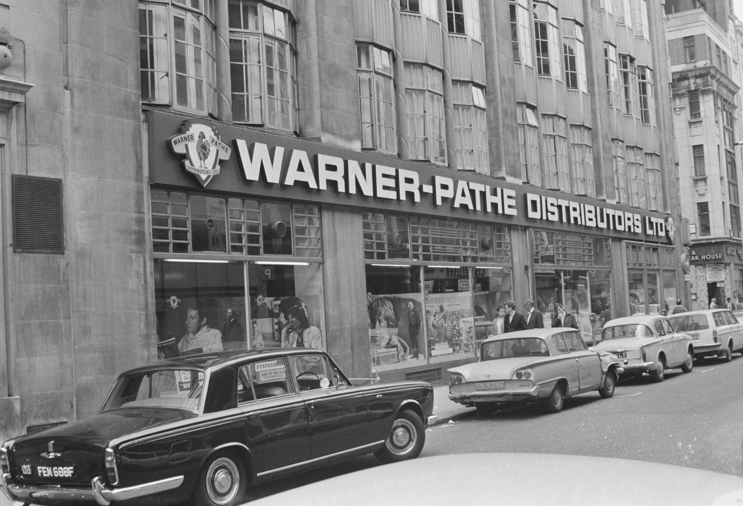 Exterior view of the Warner-Pathe Distributors office in Wardour Street, with cars parked along the street and people visible through the large windows.