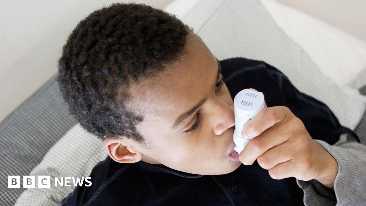 A boy is sat on a sofa or bed breathing on an inhaler.  The photo is cropped at his head and shoulders.