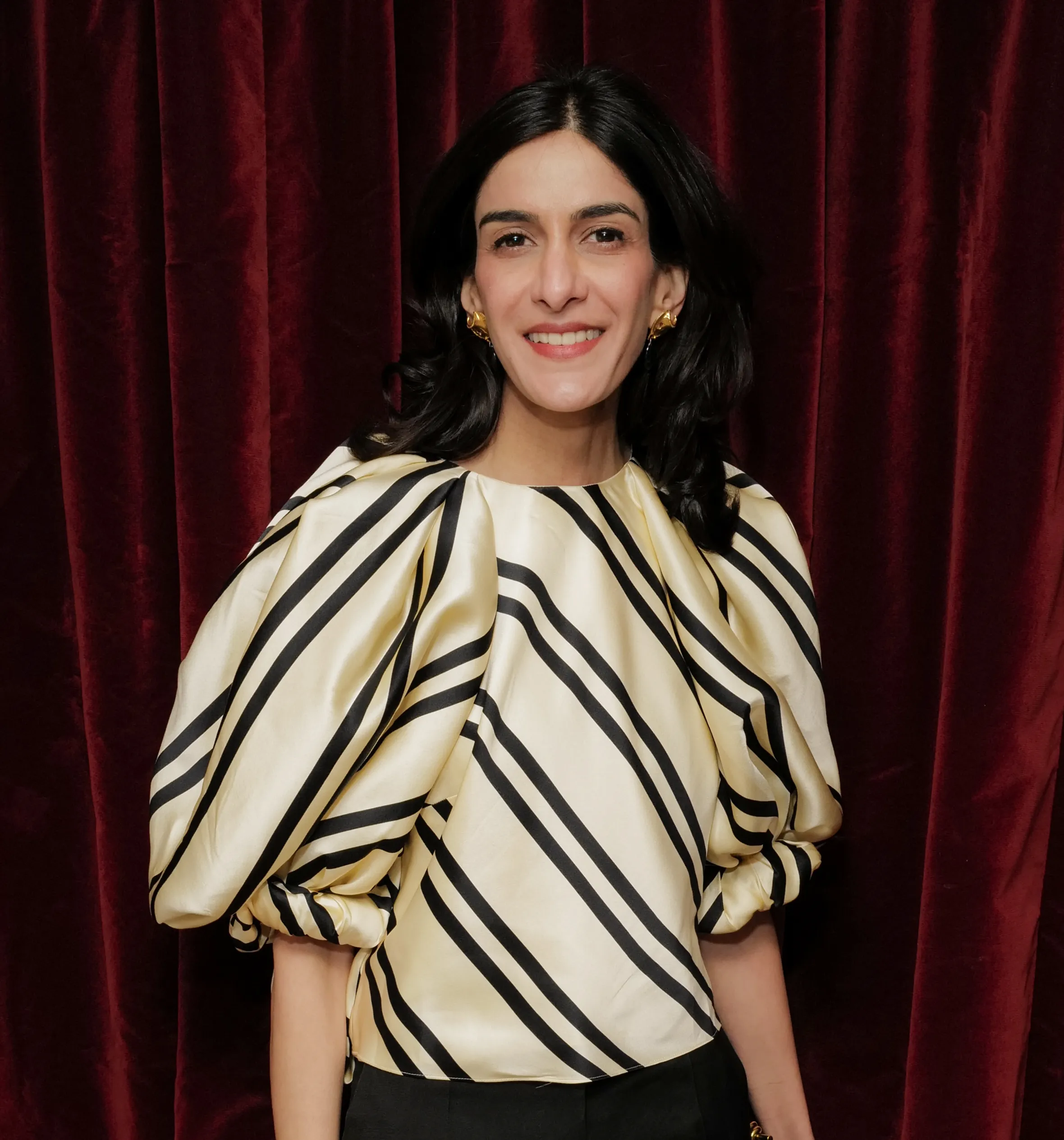 Priya Dogra in a striped blouse and black pants smiling against a red curtain.