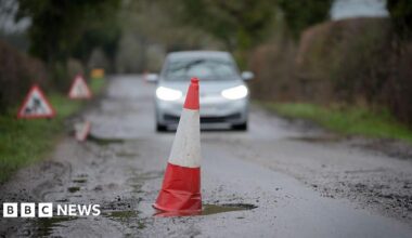 A red and white cone sits in a deep pothole. A car can be seen approaching in the background.