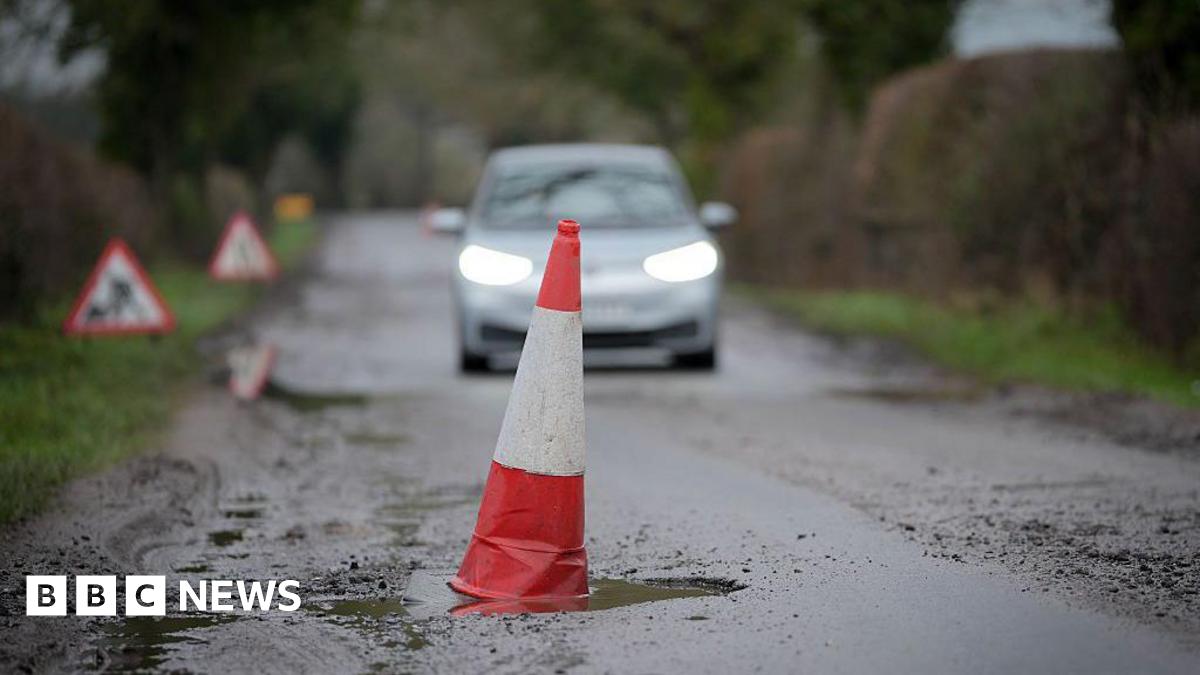 A red and white cone sits in a deep pothole. A car can be seen approaching in the background.