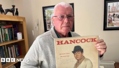 Alan Florence, has very short cropped grey hair he has silver framed glasses and is wearing a beige woollen cardigan with a green shirt beneath with the top buttons undone, behind him is a bookcase with vinyl albums and books and a small telescope in front of it.