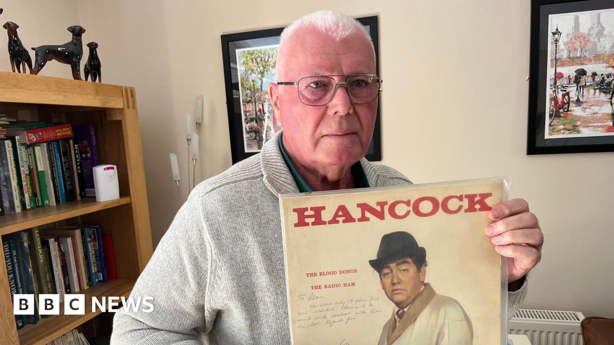 Alan Florence, has very short cropped grey hair he has silver framed glasses and is wearing a beige woollen cardigan with a green shirt beneath with the top buttons undone, behind him is a bookcase with vinyl albums and books and a small telescope in front of it.