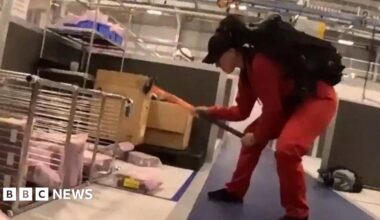 A person wearing a red jumpsuit and black cap, using a sledgehammer to damage equipment inside the factory.