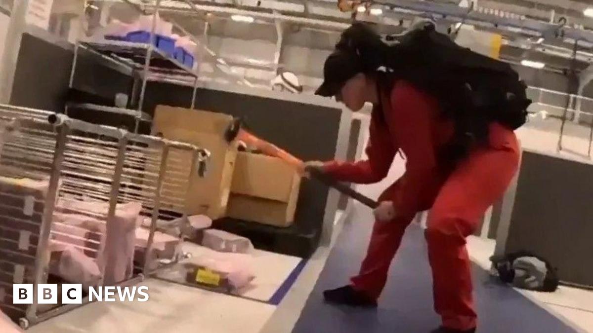 A person wearing a red jumpsuit and black cap, using a sledgehammer to damage equipment inside the factory.