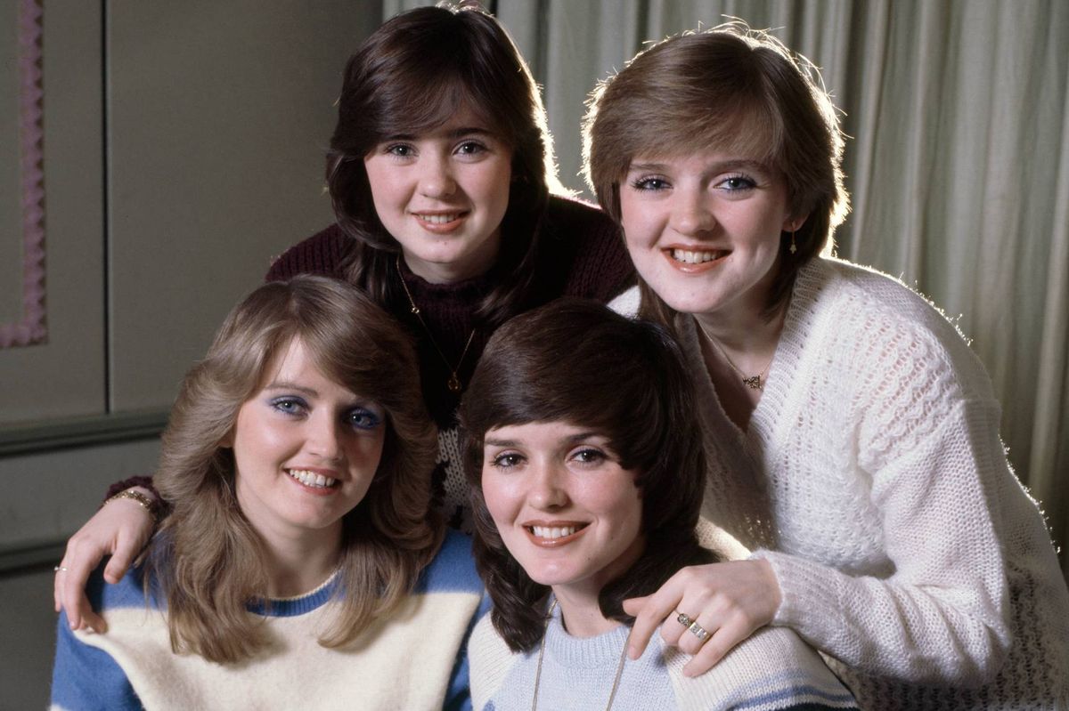 A group of four women are posing for a photograph. Three of them have their arms around each other, while the woman on the left has her arm on the shoulder of the woman in the center. They all appear to be smiling and are likely members of a musical band.