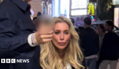 A CCTV image showing a tall man with brown hair wearing wearing a smart dark blue jacket, gesturing forward. He is with a woman with long, blonde hair