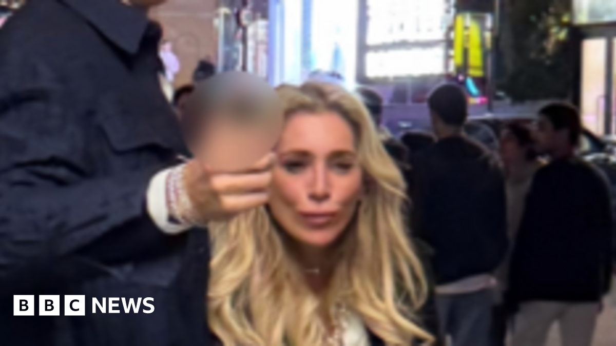 A CCTV image showing a tall man with brown hair wearing wearing a smart dark blue jacket, gesturing forward. He is with a woman with long, blonde hair